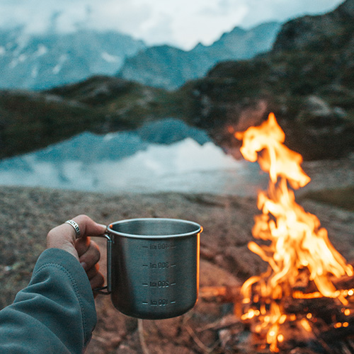 TheCalmBase | Natur – Inmitten einer Berglandschaft brennt beim Camping ein Lagerfeuer. Eine Hand hält eine Tasse in Richtung des Feuers.