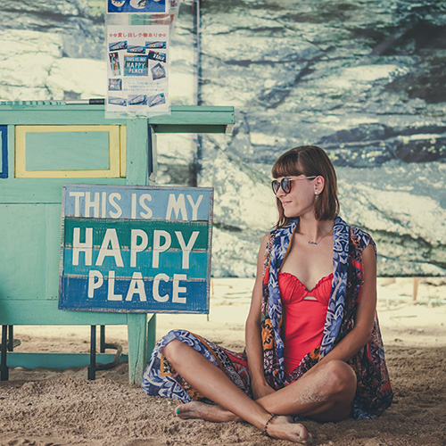 TheCalmBase | Wohlbefinden – Eine Frau sitzt entspannt im Sand neben einem Schild auf dem steht: This is my happy place