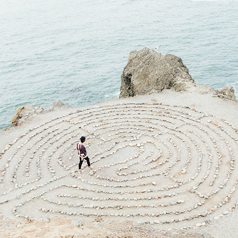 TheCalmBase | Meditation – Eine Person geht am Strand durch ein Labyrinth aus Steinen. Im Hintergrund rauscht das Meer.