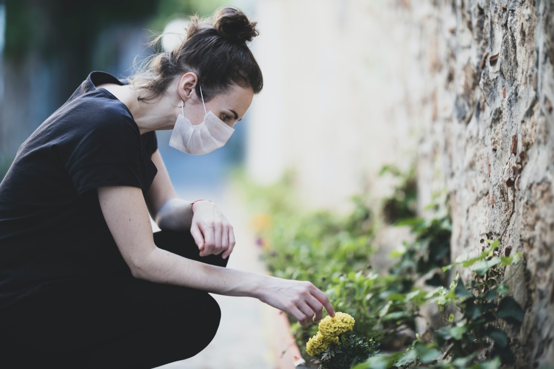 Eine Frau trägt eine Maske und erfreut sich an einer kleinen Blume am Straßenrand.