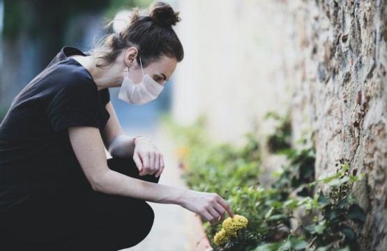 Eine Frau trägt eine Maske und erfreut sich an einer kleinen Blume am Straßenrand.